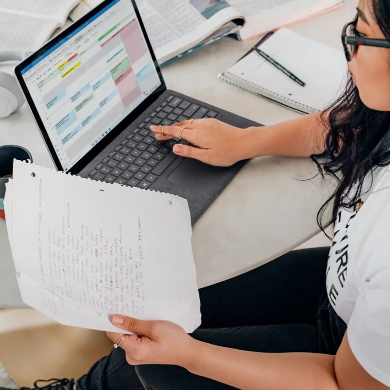 A person at a scattered desk surrounded by open journals, hand-written notes, and a laptop calendar — searching across multiple sources to find a single answer.